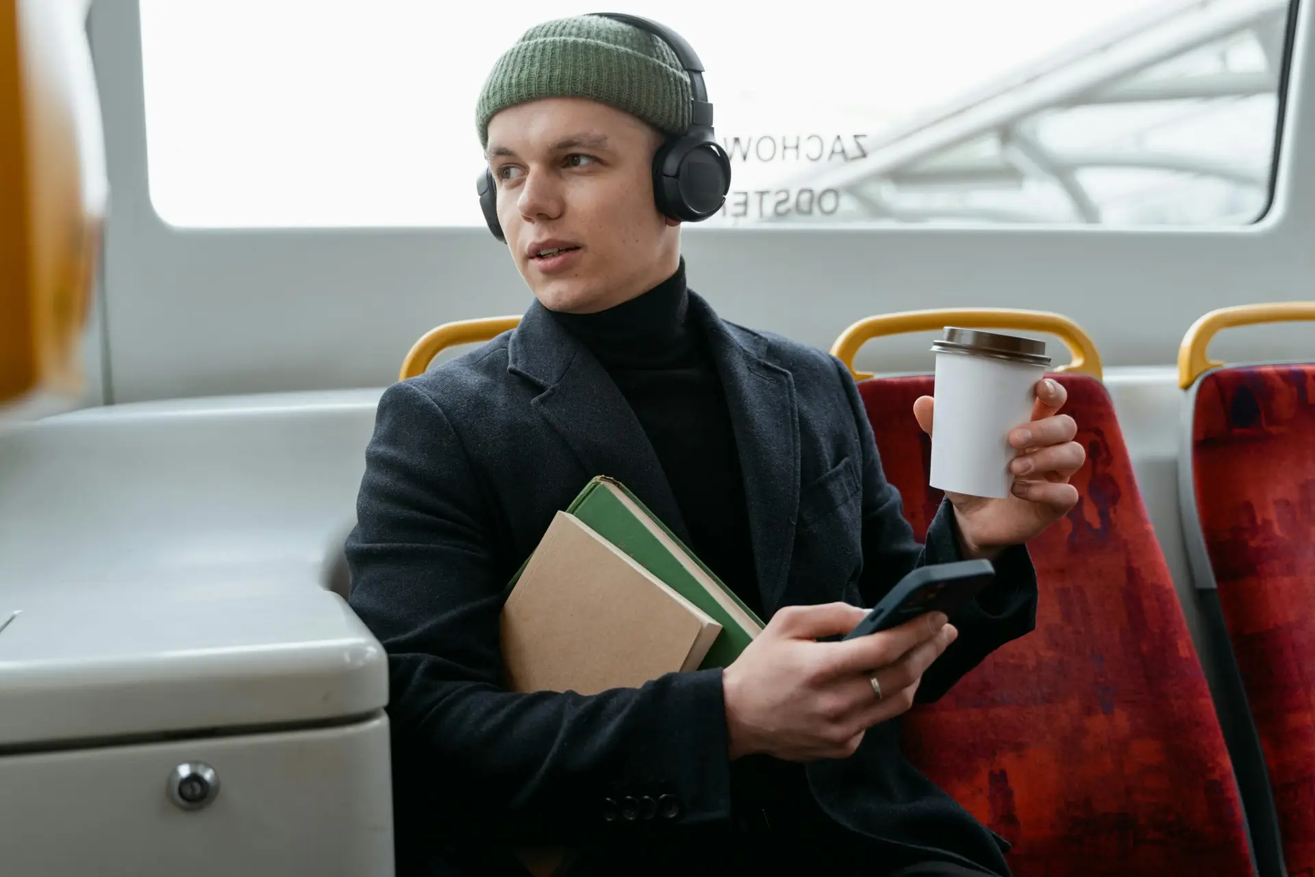 Man wearing headphones, holding coffee on a bus, using cellphone.
