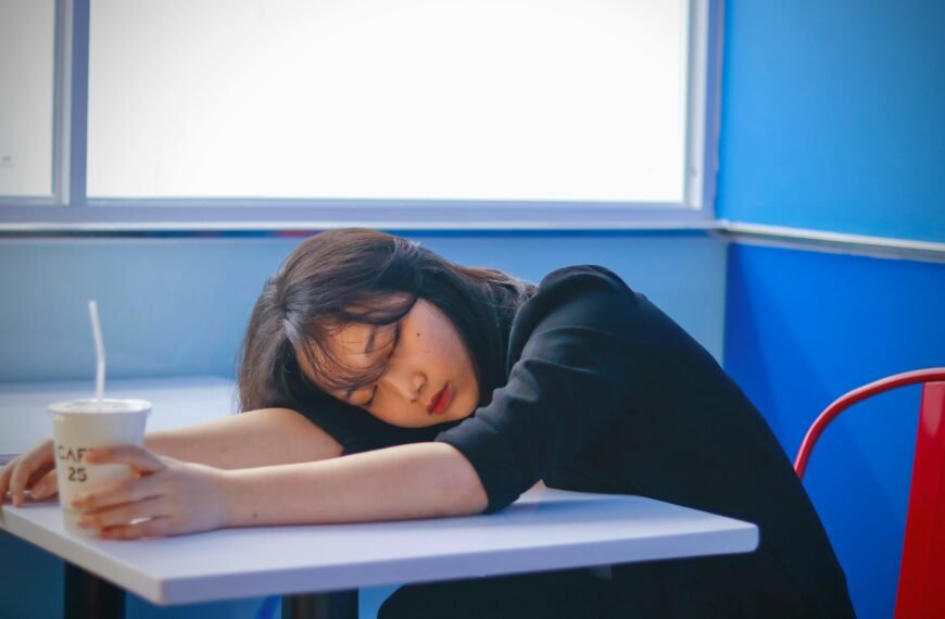 A woman sleeping on a café table, holding a disposable coffee cup, looking tired.